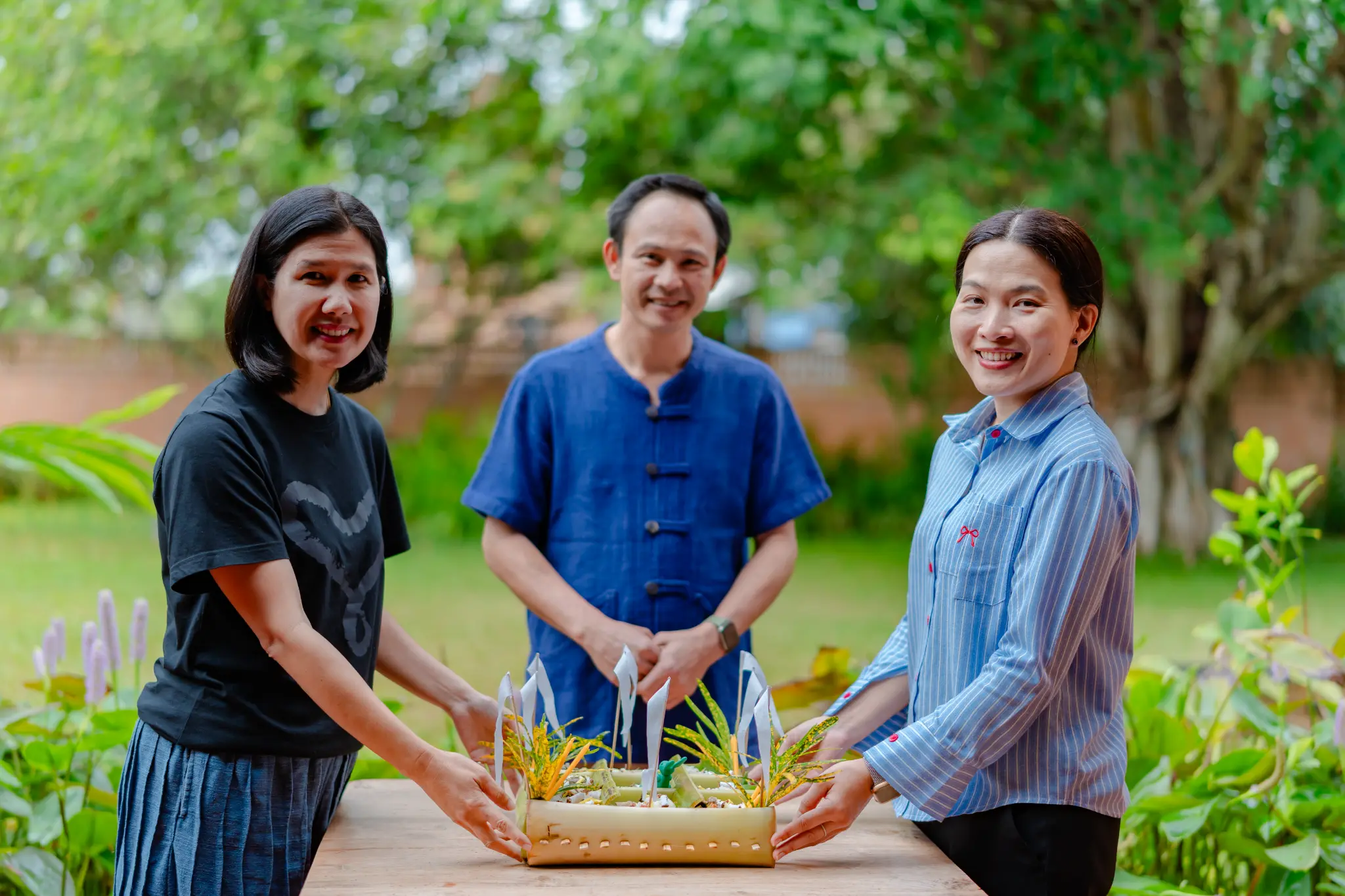 Three people holding a handmade bamboo piece with green and yellow plant material
