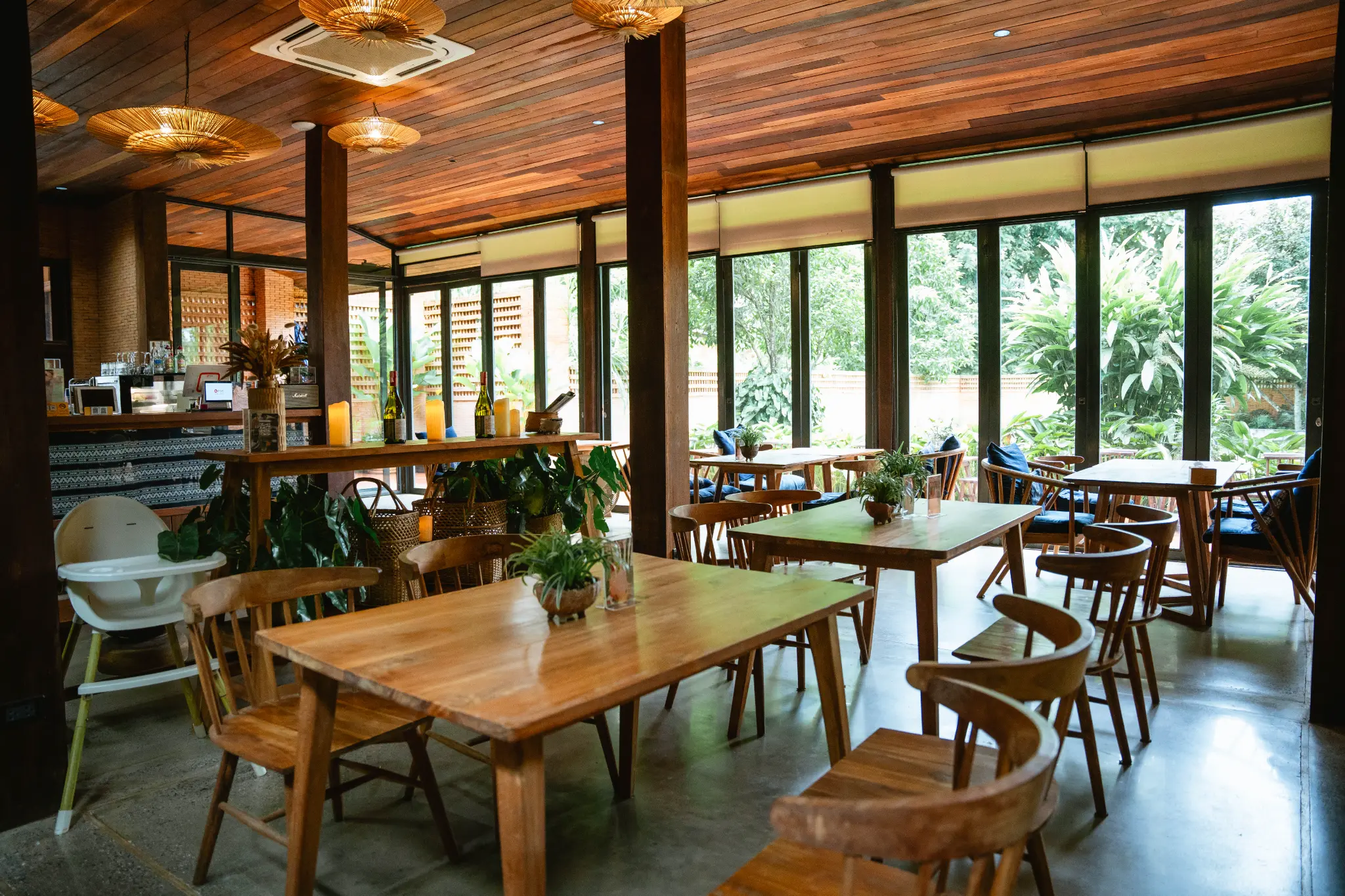 Dining room facing the garden windows. A bright dining room with wooden furniture faces large windows and dense green garden views. Photographed at Wiang Neua Café & Restaurant in Chiang Saen, Chiang Rai.