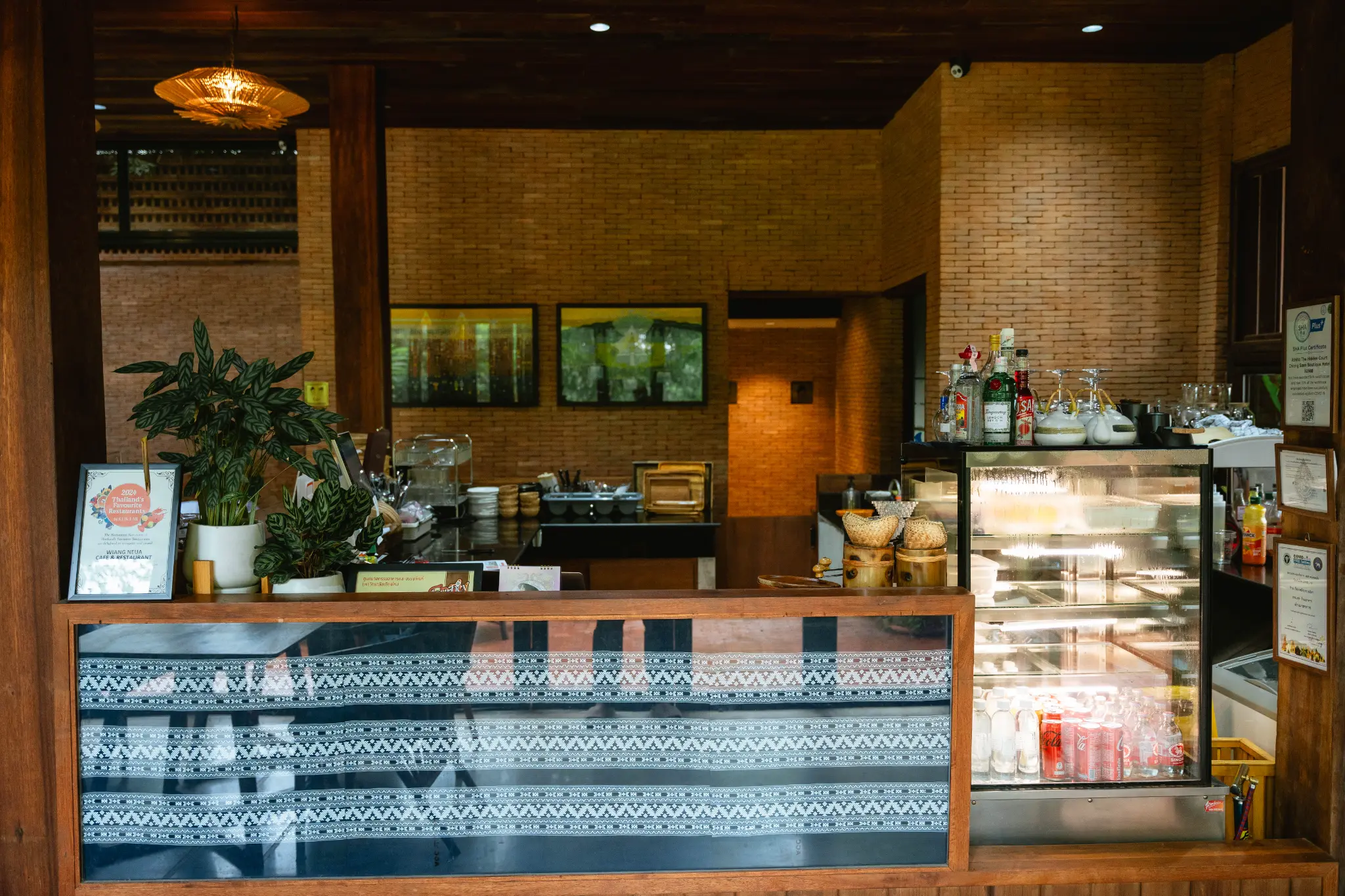 Café counter against old brick. A café counter with a display cabinet, bottles and plants stands against a warm brick wall. Photographed at Wiang Neua Café & Restaurant in Chiang Saen, Chiang Rai.