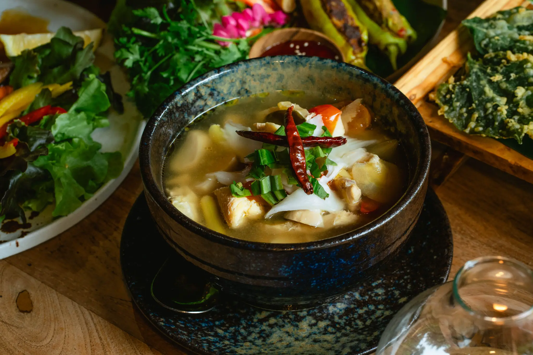 Clear soup with vegetables and side dishes. A clear soup with mushrooms, vegetables, dried red chilies and green onions is accompanied by salad and fried greens. Photographed at Wiang Neua Café & Restaurant in Chiang Saen, Chiang Rai.