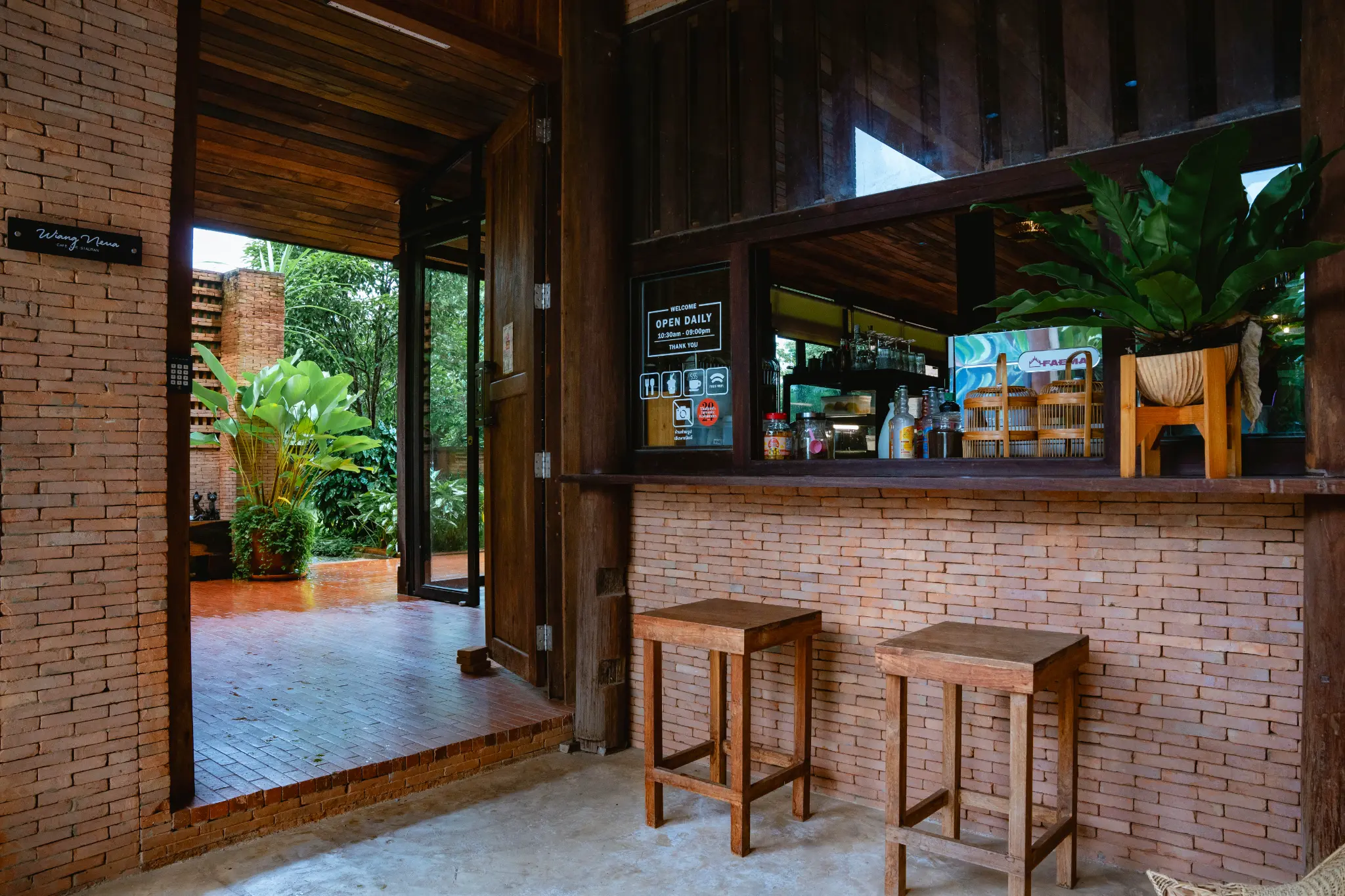 Brick counter and garden doorway. A brick counter with wooden stools, bottles, jars and plants opens toward a green garden view. Photographed at Wiang Neua Café & Restaurant in Chiang Saen, Chiang Rai.