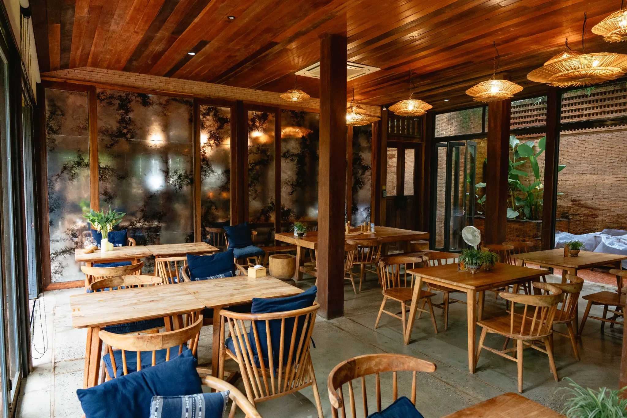Intimate dining corner with blue cushions. An intimate wooden dining corner has blue-cushioned chairs, woven lights and glass walls reflecting the room. Photographed at Wiang Neua Café & Restaurant in Chiang Saen, Chiang Rai.