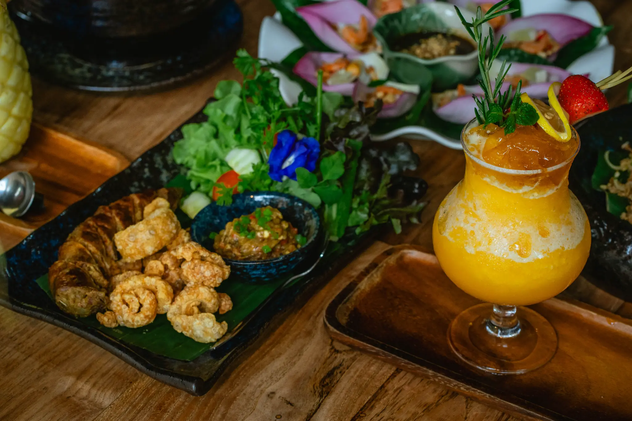Fried bites, fresh greens and a citrus-coloured drink. Fried bites, fresh greens, a small bowl of dip and an orange-coloured garnished drink are arranged on a wooden table. Photographed at Wiang Neua Café & Restaurant in Chiang Saen, Chiang Rai.