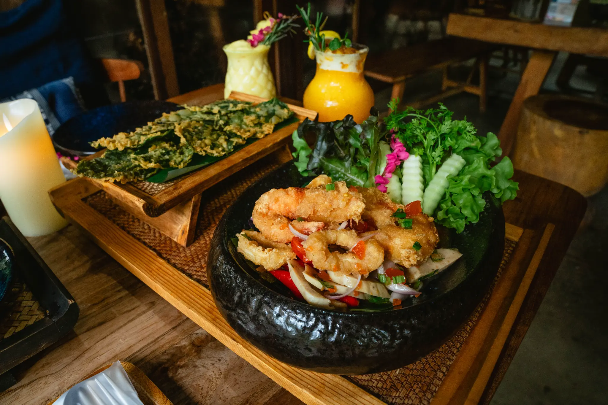 Fried shrimp and fresh greens. Fried shrimp, fresh salad, fried leafy greens and two yellow drinks are presented on a wooden table. Photographed at Wiang Neua Café & Restaurant in Chiang Saen, Chiang Rai.