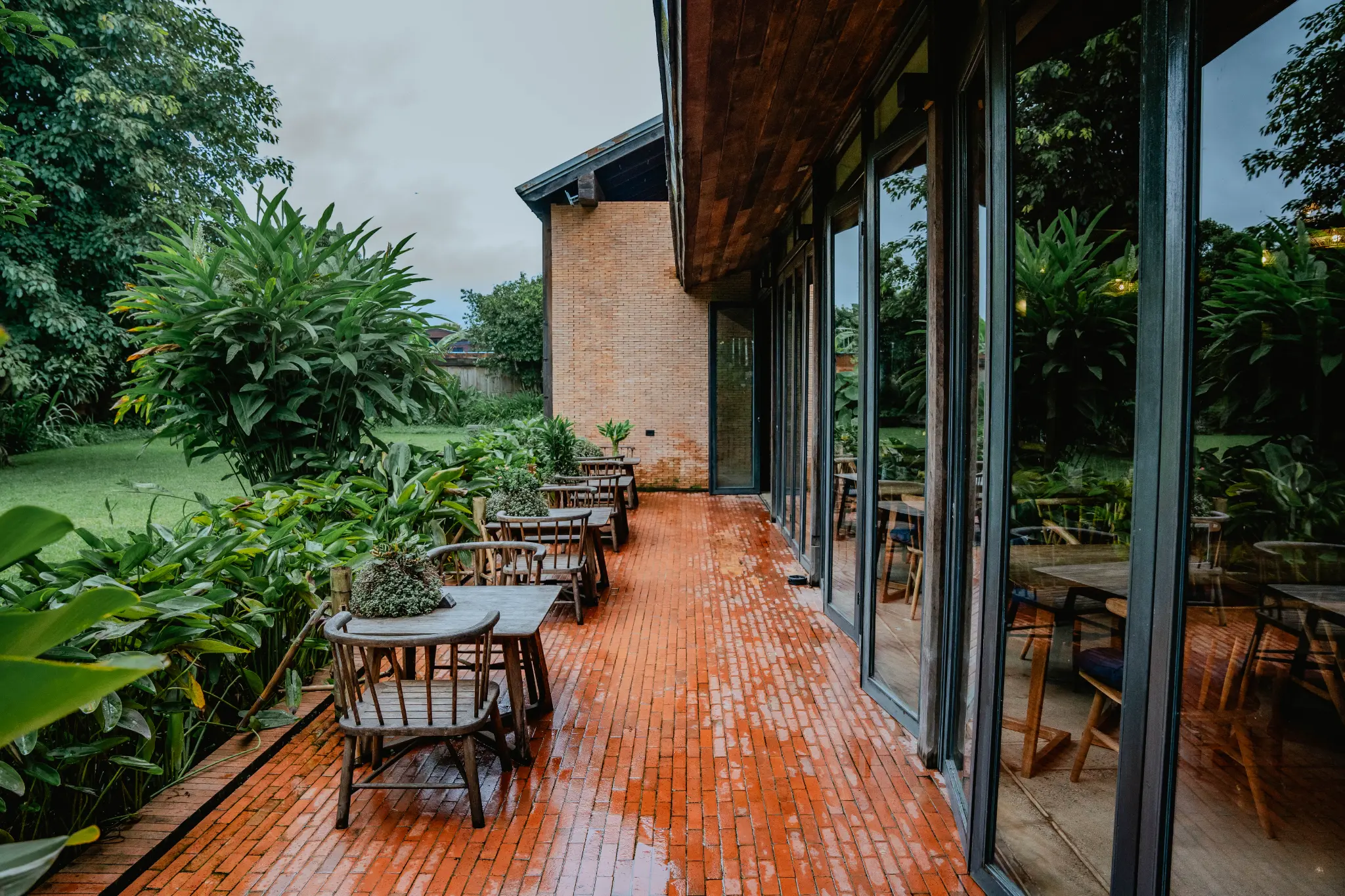 Tables along the garden-side brick walk. Wooden tables line a wet red-brick walkway between reflective windows and dense garden planting. Photographed at Wiang Neua Café & Restaurant in Chiang Saen, Chiang Rai.