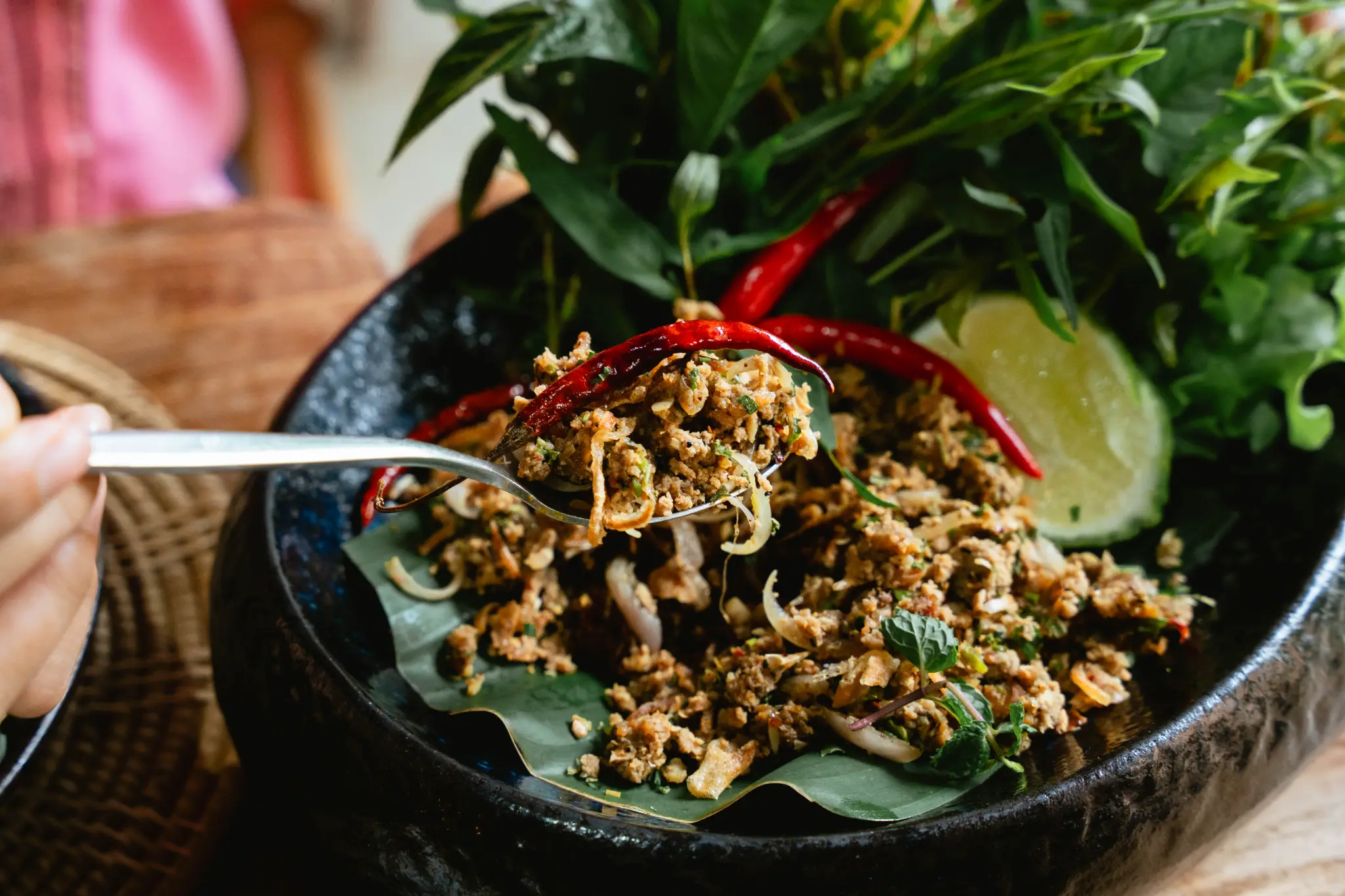Minced dish with herbs, chili and lime. A minced dish is garnished with fresh herbs, red chili and lime in a dark bowl as a spoon lifts one portion. Photographed at Wiang Neua Café & Restaurant in Chiang Saen, Chiang Rai.