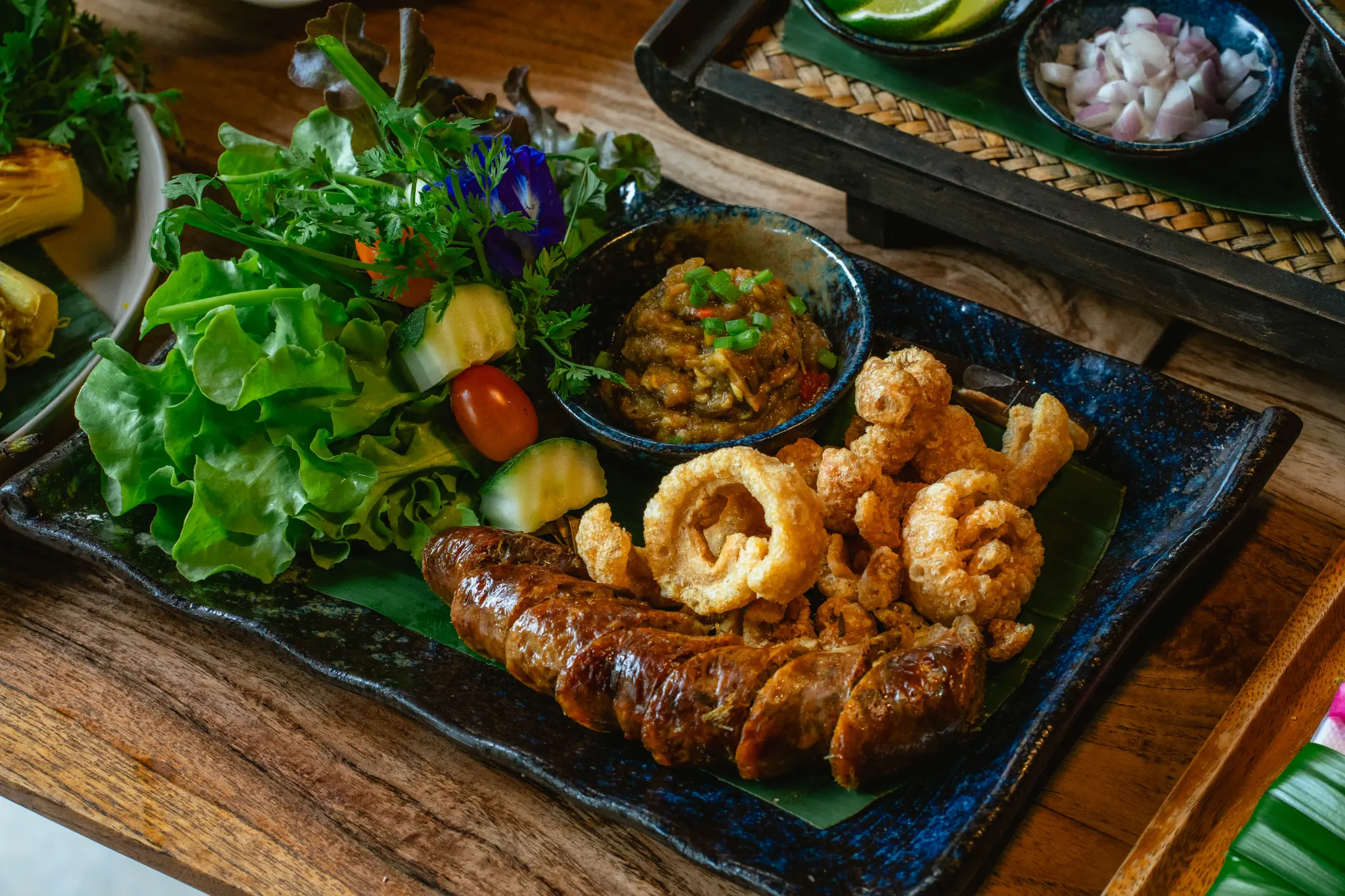 Northern-style sharing platter. Sliced grilled sausage, crispy pork rind, a green dip, cucumber, tomato, lettuce and herbs are arranged on a dark platter. Photographed at Wiang Neua Café & Restaurant in Chiang Saen, Chiang Rai.