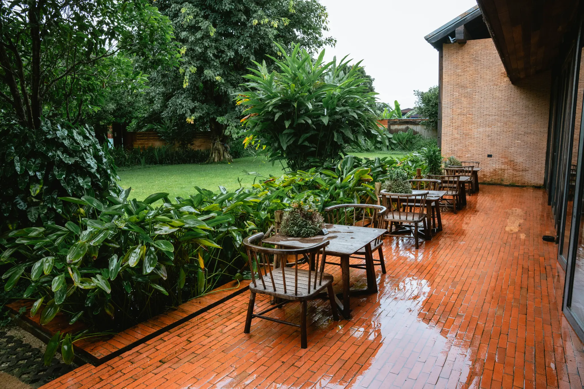 Garden-side outdoor dining. Wooden tables and chairs are arranged on a red-brick patio surrounded by dense green plants. Photographed at Wiang Neua Café & Restaurant in Chiang Saen, Chiang Rai.