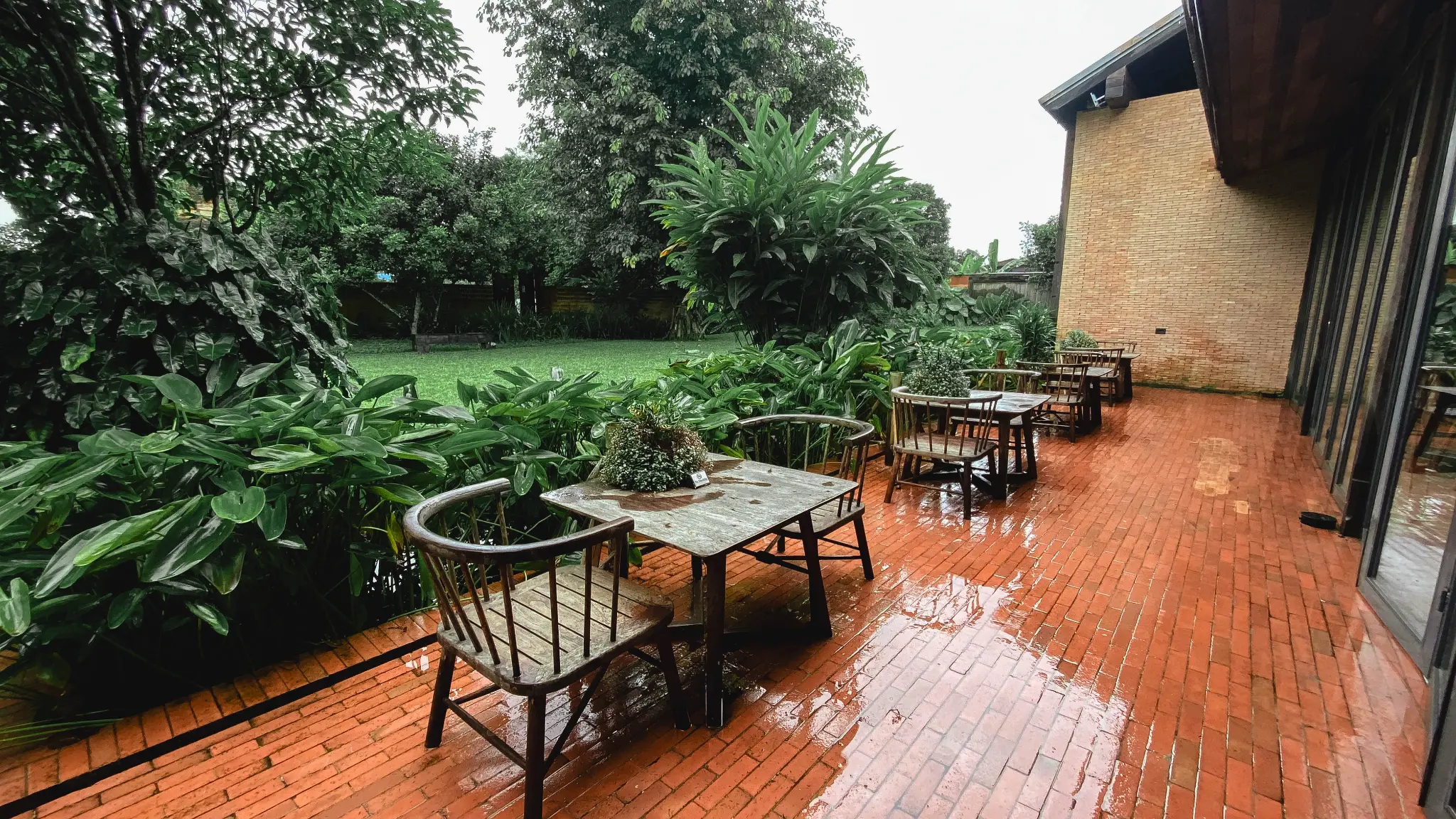 Wide garden patio dining. A wide view shows several wooden tables across a red-brick patio beside a green garden and brick building. Photographed at Wiang Neua Café & Restaurant in Chiang Saen, Chiang Rai.