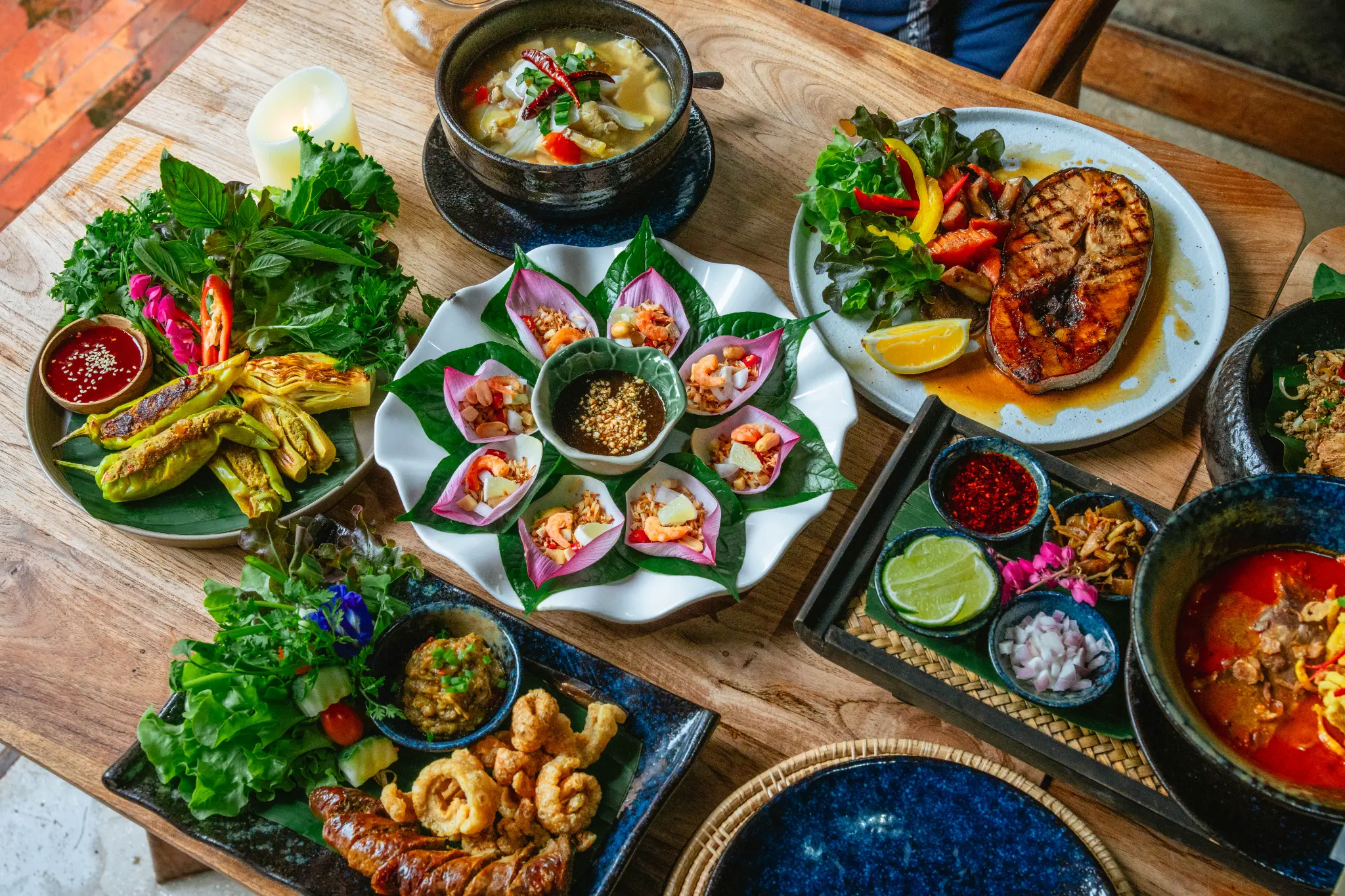Overhead view of a shared table. An overhead composition shows several colourful dishes arranged across a wooden dining table. Photographed at Wiang Neua Café & Restaurant in Chiang Saen, Chiang Rai.
