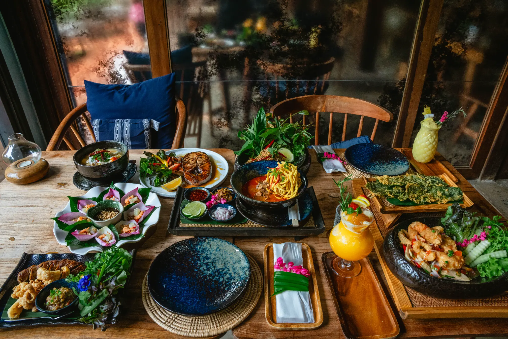 Shared table beside the window. Multiple dishes, place settings and two drinks fill a wooden table beside a window and blue-cushioned chairs. Photographed at Wiang Neua Café & Restaurant in Chiang Saen, Chiang Rai.