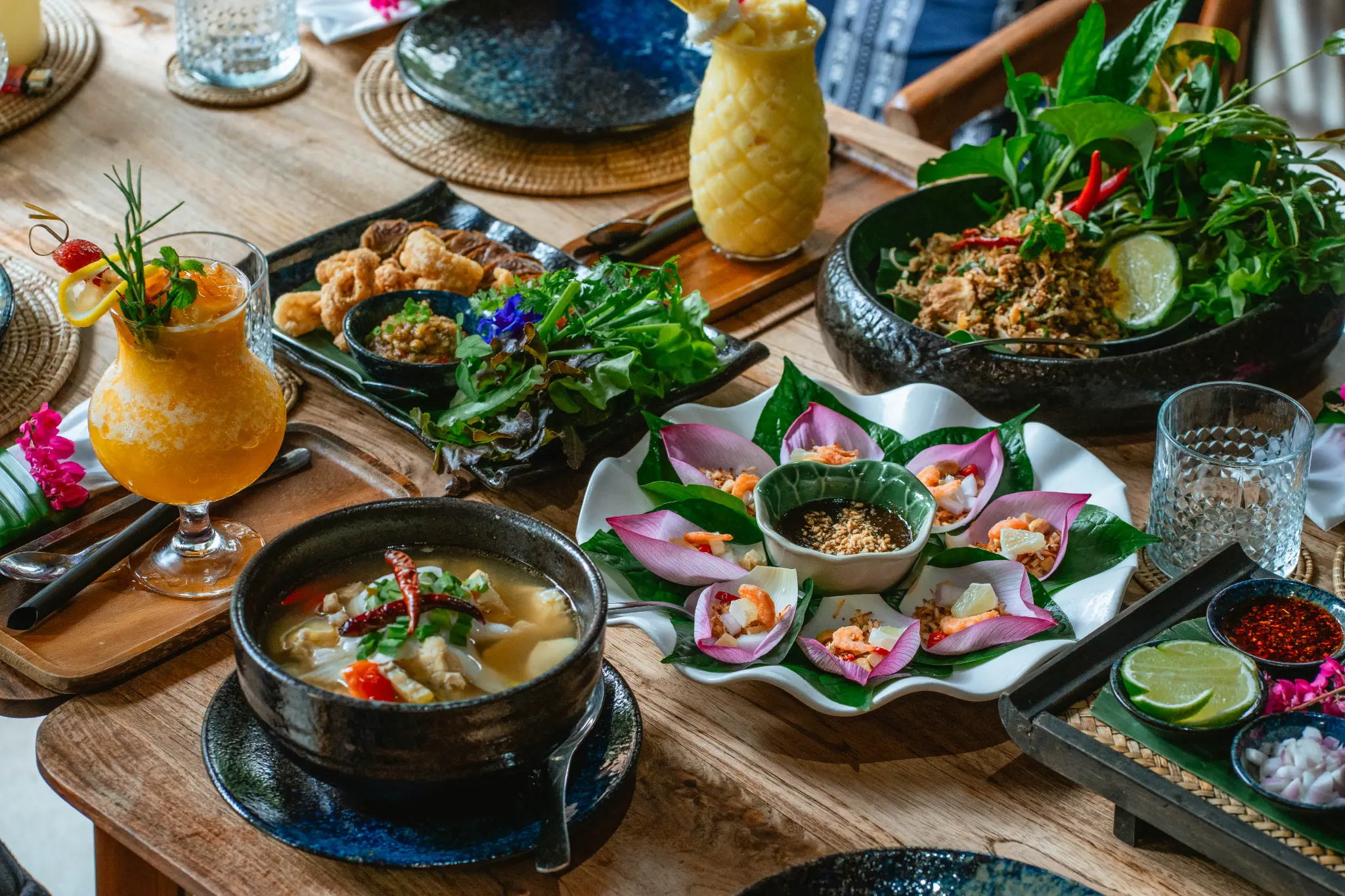 A shared table of dishes. Several dishes, a bowl of soup, fried items, fresh vegetables and two blended drinks share a wooden table. Photographed at Wiang Neua Café & Restaurant in Chiang Saen, Chiang Rai.