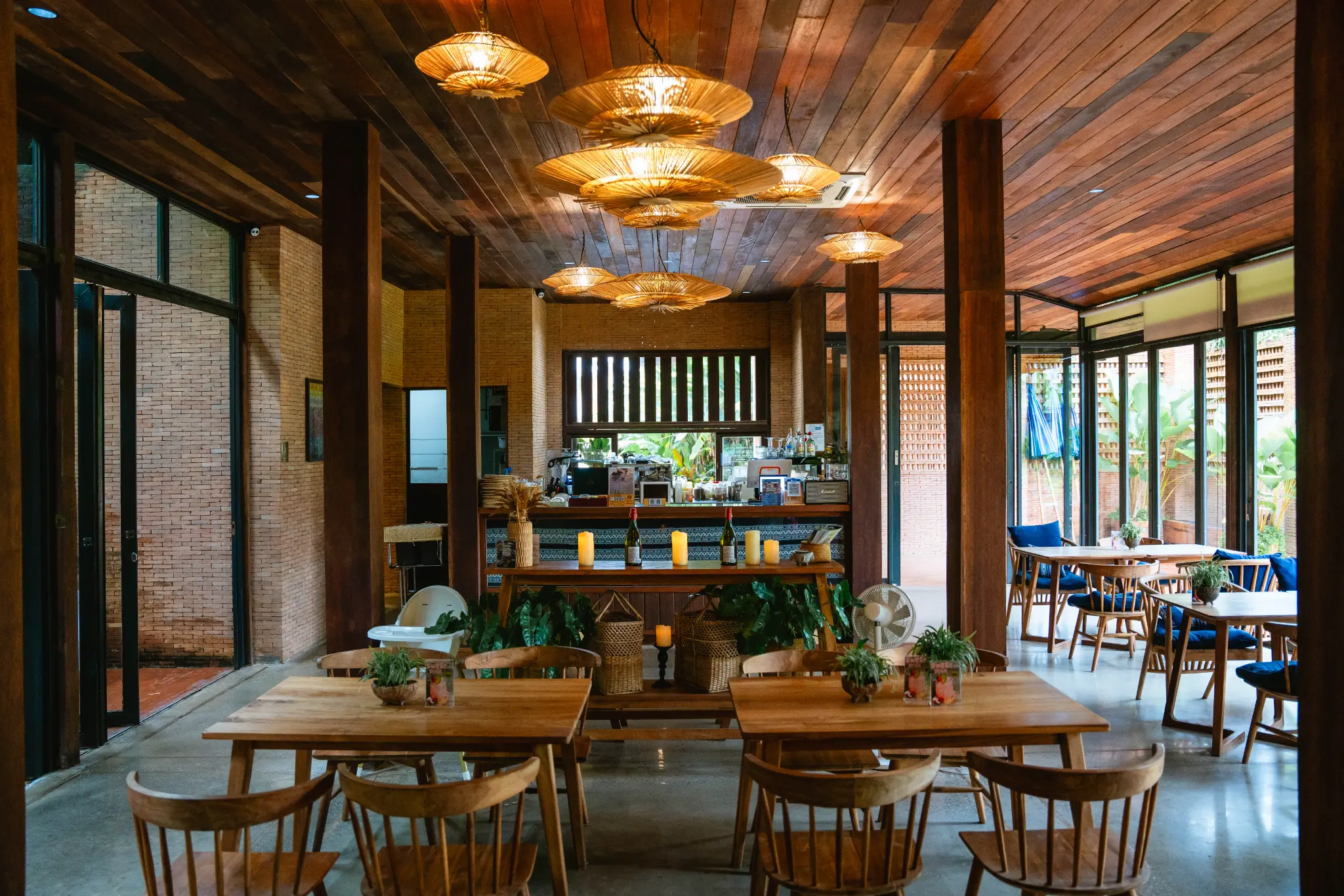 Wide view of the wooden dining room. Wooden tables and chairs sit beneath woven pendant lights with a service counter at the far end. Photographed at Wiang Neua Café & Restaurant in Chiang Saen, Chiang Rai.