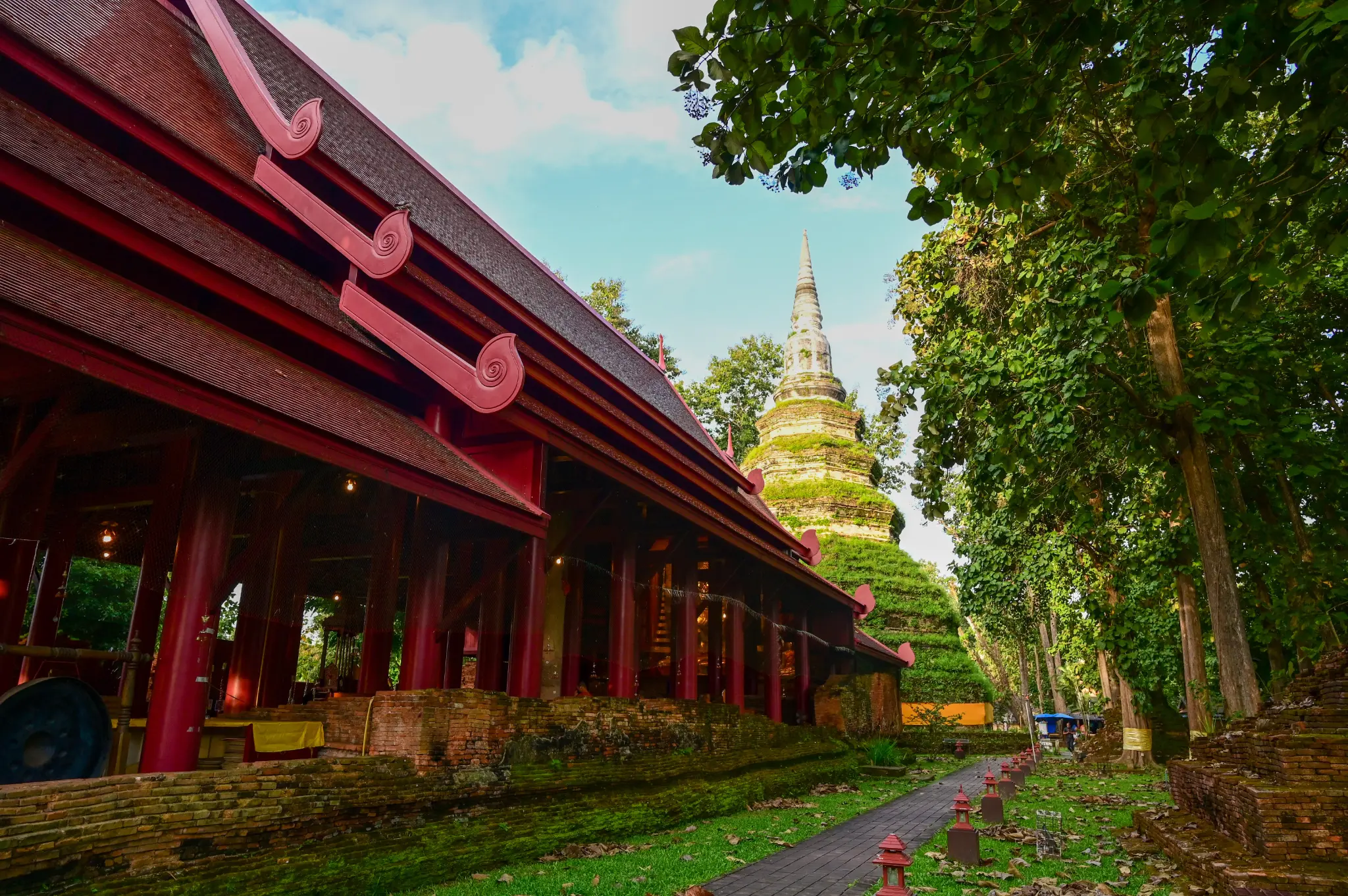 A red-roofed Thai building, ancient stupa and brick path among green trees